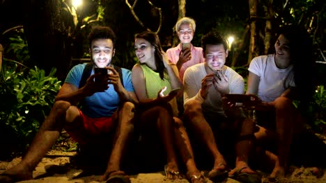 Grupo-de-personas-usando-Smart-teléfono-celular-risa-sentado-sobre-un-banco-en-el-parque-de-noche,-jóvenes-amigos,-hablando-de-redes-en-línea