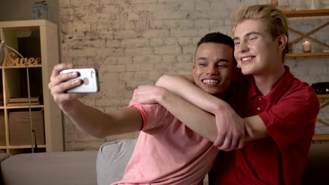 Two-international-gay-friends,-sitting-on-the-couch,-hugging,-making-selfies,-laughing.-Homeliness,-romantic-evening,-background,-hugs,-happy-LGBT-family-concept-60-fps