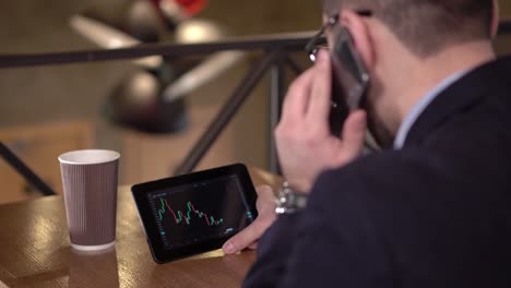 Young-man-monitors-trading-cryptocurrency-on-tablet-and-speaks-phone-in-a-cafe