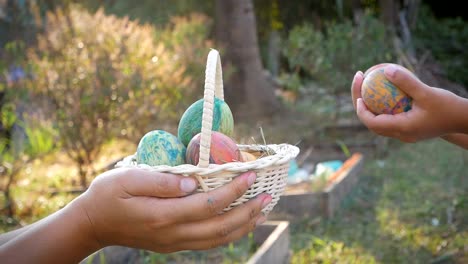 Primer-plano-de-mano-de-mujer-sosteniendo-una-cesta-con-huevos-de-Pascua-y-su-dando-a-los-niños-en-el-fondo-de-la-luz-del-sol