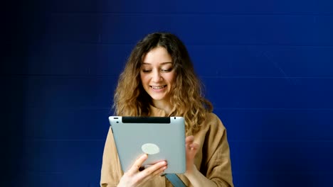 Young-woman-uses-a-tablet-for-video-communication-on-a-blue-wall-background