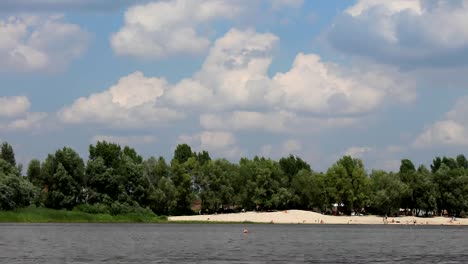 Menschen-am-Strand-des-gegenüberliegenden-Flussufer-unter-blauem-Himmel-mit-flauschigen-Wolken.-Zeitraffer