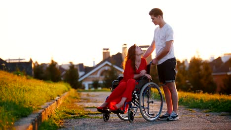 Girl-In-A-Wheelchair-Cute-Talking-To-Her-Young-Man-On-The-Background-Of-The-House,-Green-Grass-And-Trees