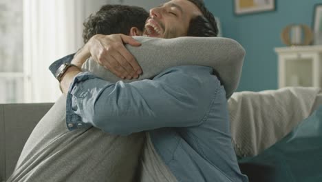 Pareja-Gay-masculino-atractivo-lindo-sentarse-juntos-en-un-sofá-en-casa.-Novios-son-Hugging-y-abrazándose-unos-a-otros.-Ellos-son-feliz-y-sonriente.-Son-vestidos-casualmente-y-sala-tiene-un-Interior-moderno.