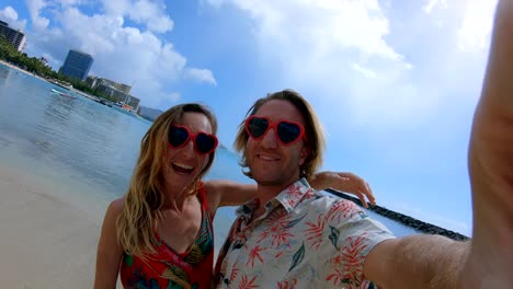 Couple-taking-selfies-on-Waikiki-Beach-in-Honolulu,-Hawaii.-Selfie-point-of-view-wide-angle-of-Waikiki-Beach.-Young-couple-taking-selfies-with-heart-shaped-sunglasses