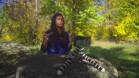 Blogging-young-woman-sitting-next-to-camera-on-the-nature-talking-and-gesturing-and-smiling