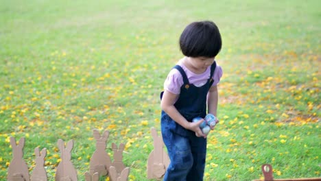 Niedliches-kleines-Kindermädchen-am-Ostersamstag.-Mädchen-jagt-nach-Ostereiern-auf-dem-Rasen-und-Hase-Made-of-paper-in-der-Natur-oder-Park-und-Sonnenlicht.-Zeitlupe