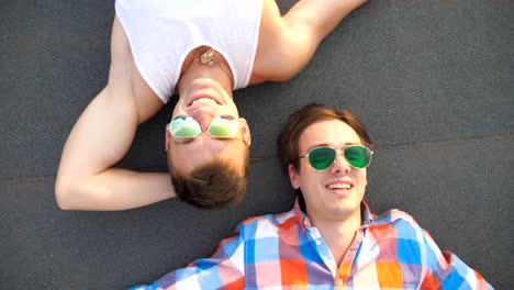 Joven-pareja-masculina-feliz-en-las-gafas-de-sol-tumbado-en-la-azotea-del-edificio-de-gran-altura-y-la-risa-franca.-Guapos-chicos-gays-mirando-cámara-y-sonriendo-mientras-hablan-en-el-techo.-Vista-superior-cerrar-cámara-lenta