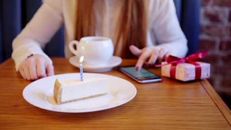 Close-up-of-waitress-serving-piece-of-birthday-cake-with-burning-candle-for-woman-having-tea-and-browsing-smartphone-alone-in-cafe,-waiting-for-somebody-to-come.-Birthday-gift-box-placed-on-table