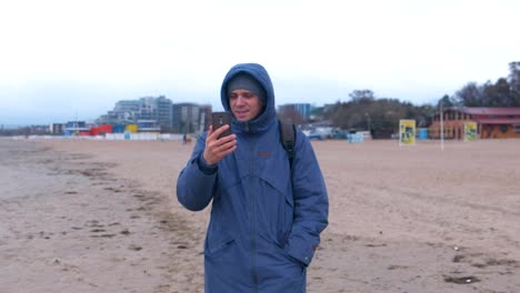 Mann-Blogger-in-einer-blauen-Daunenjacke-zu-Fuß-auf-dem-Sandstrand-am-Meer-und-sprechen-einen-Video-Chat-auf-Handy.