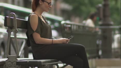 Attractive-redhead-woman-with-glasses,-freckles,-piercings-and-red-hair-writing-a-text-message-on-her-smartphone-sitting-on-street-bench,-during-sunny-summer-in-Paris.-Tilt-up-Slow-motion.-Trendy.