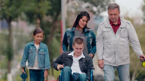 Happy-parents-walk-with-children.-Son-in-a-wheelchair-for-a-walk-with-mom,-dad,-sister-and-dog.-Portrait-view.