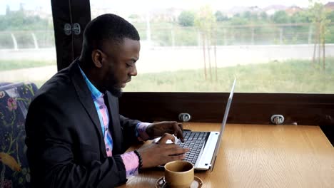 Black-businessman-is-working-typing-a-message-on-laptop-sitting-in-summer-cafe.