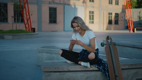 Beautiful-Blonde-Skater-Girl-Wearing-Casual-Clothes,-Uses-Smartphone,-Sharing-Stuff-on-Social-Media,-Taking-Selfies,-Messaging-Friends-and-Having-Fun-while-Sitting-on-a-Bench-in-Fashionable-Hipster