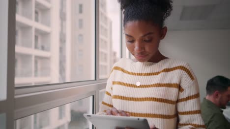 Porträt-einer-lächelnden,-trendigen-afrikanischen-jungen-Frau-mit-digitalem-Tablet,-die-vor-seinem-Kollegen-im-Hintergrund-sitzt