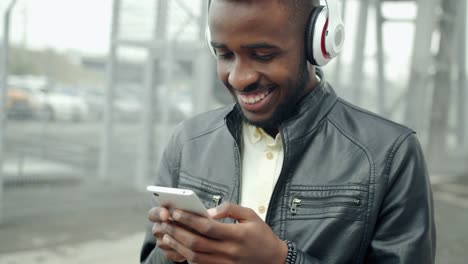 African-American-student-in-headphones-touching-smartphone-screen-outdoors