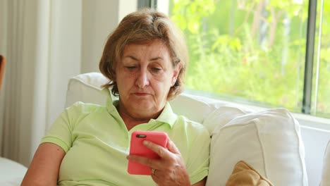 Thoughtful-candid-older-senior-woman-holding-smartphone-device-seated-at-home-sofa