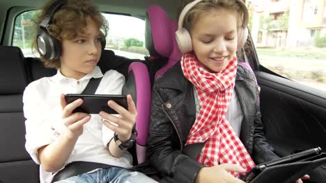 Boy-and-Girl-with-Headphones-Playing-a-Tablet-and-smartphone-in-a-Car,-Children-Using-a-Devices-in-the-auto.-Brother-and-Sister-Traveling-Together