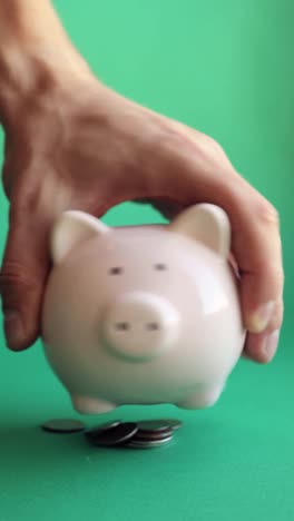 Man-shaking-piggy-bank-and-coins-fall-out-on-green-background,-vertical-video