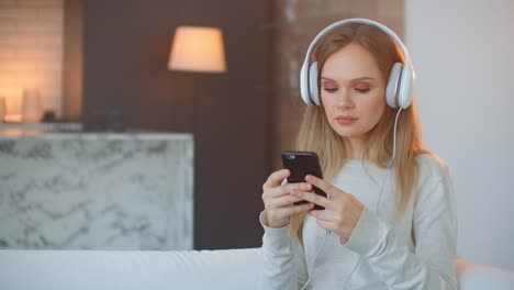 Calma-feliz-joven-mujer-en-auriculares-escalofriante-sentado-en-el-sofá-con-los-ojos-cerrados-escuchando-música-favorita-sosteniendo-el-teléfono-usando-la-aplicación-reproductor-en-línea-móvil-disfrutar-de-estado-de-ánimo-pacífico-usando-auriculare