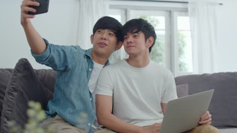 Romantic-young-gay-couple-funny-selfie-by-cellphone-at-home.-Asian-lover-male-happy-relax-fun-using-technology-mobile-phone-smiling-take-a-photo-together-while-lying-sofa-in-living-room-concept.