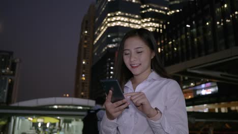 Joven-mujer-asiática-sonriente-de-negocios-usando-teléfono-inteligente-en-el-centro-de-la-ciudad-moderna-en-la-noche-de-Bangkok-Tailandia.-Comunicación-Concept-Technology-por-teléfono-móvil