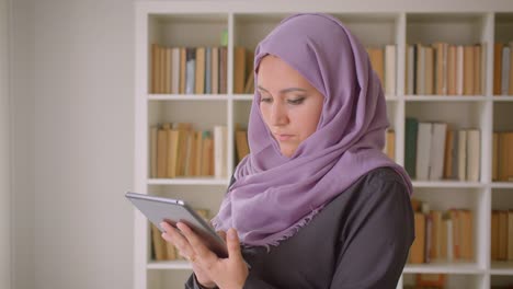 Retrato-de-primer-plano-de-joven-mujer-musulmana-en-hiyab-usando-la-tableta-y-mirando-la-cámara-de-pie-en-la-biblioteca-en-el-interior