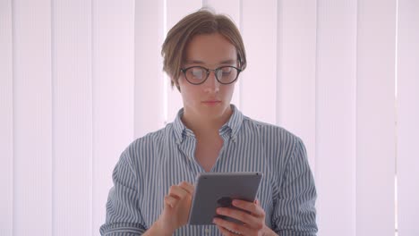 Closeup-portrait-of-young-handsome-caucasian-businessman-in-glasses-using-the-tablet-standing-indoors-in-the-office