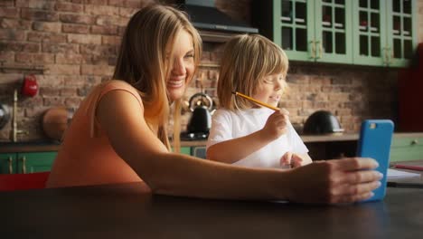 Mamá-y-niño-sonriendo-en-traje-casual-están-disfrutando-de-videollamada-en-línea-en-el-teléfono-móvil-mientras-hacen-la-tarea.-Se-sentaron-en-la-mesa-en-la-cocina.-De-cerca