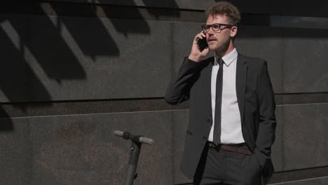 Employee-talking-on-cellphone-near-office-building,-electric-scooter-nearby