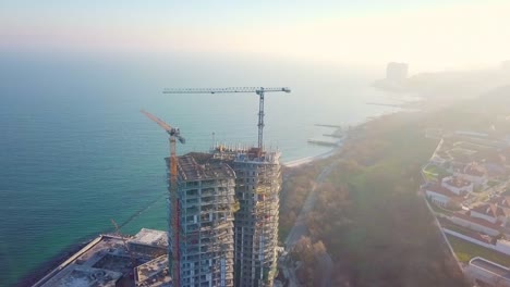 Aerial-city-view.-Construction-of-a-high-rise-skyscraper-on-the-ocean-by-two-cranes.-Around-clockwise.-The-rays-of-the-setting-sun-fall-into-the-lens