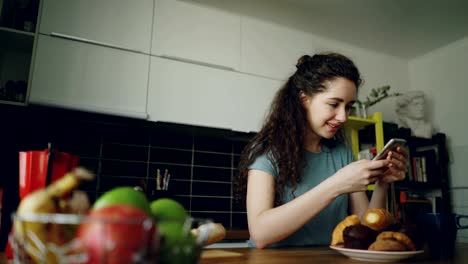 young-beautiful-curly-pretty-caucasian-woman-sitting-at-table-in-nice-kitchen-using-smartphone,-she-is-texting-someone-and-smiling,-calm-and-happy