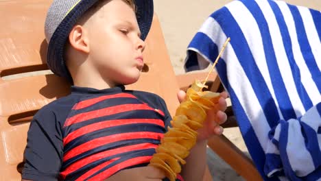 Niño-comiendo-patatas-fritas-caseras-en-un-palo,-en-la-playa-del-mar.-Pone-en-una-tumbona.