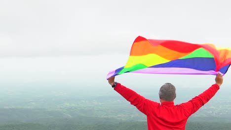 Mann-erhöhen-Regenbogen-Farbe-LGBTI-Fahnenschwingen-in-harten-Wind-auf-Mountain-Top-Sicht