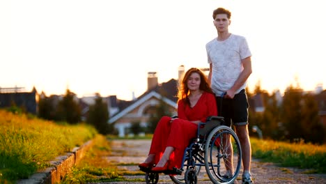 Girl-On-Wheelchair-And-Her-Boyfriend-Look-At-The-Camera-And-Smile-Slightly-In-The-Background-Of-Green