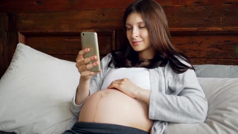 Schöne-schwangere-Mädchen-Brünette-ist-ihr-Babybauch-streicheln-und-mit-Smartphone-gemütlich-im-Bett-zu-Hause-mit-glücklichen-Lächeln.-Schwangerschaft,-Familie-und-Technologie-Konzept.