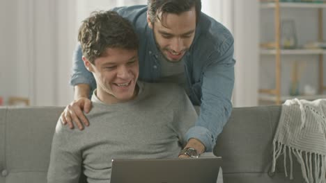 Süße-männliche-Queer-paar-verbringen-Zeit-zu-Hause.-Junger-Mann-verwendet-einen-Laptop,-seine-Partnerin-von-hinten-kommt-und-sanft-umarmt-ihn.-Sie-lachen-und-Hände-berühren.-Zimmer-ist-Modern-eingerichtet.