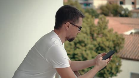 Male-user-is-reading-messages-in-smartphone,-swiping-screen,-standing-in-balcony