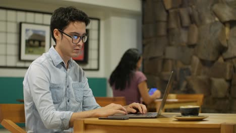 Hombre-hispano-sentado-en-la-cafetería-escribiendo-en-un-ordenador-portátil,-chica-en-el-fondo