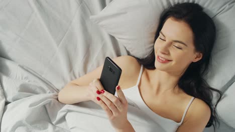 Portrait-of-Beautiful-Brunette-Using-Touchscreen-Smartphone-in-Her-Bed,-Chatting-with-Friends,-Using-Social-Media,-Posting-Pictures,-Sharing-Thoughts.-Top-Down-Camera-Shot.