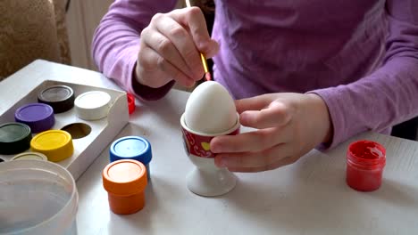 A-little-girl-draws-with-a-brush-and-colored-paints-on-a-white-egg,-sitting-at-home-at-the-table.-Close-up.-4K.