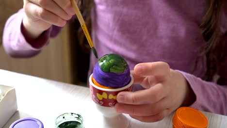 A-little-girl-paints-an-Easter-egg-with-colorful-colors-sitting-at-home-at-the-table.-Close-up.-4K.