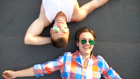 Vista-superior-de-los-chicos-gays-guapos-en-las-gafas-de-sol-tumbados-en-la-azotea-del-edificio-de-gran-altura-y-riendo.-Joven-pareja-masculina-feliz-mirando-a-la-cámara-y-mostrando-diferentes-grimases-en-el-techo.-Cerrar-cámara-lenta