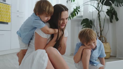 Loving-mother-and-children-hugging-and-having-fun-playing-sitting-on-the-floor.-Fun-and-happy-family.-The-game-after-viewing-the-laptop