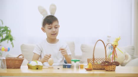 Cute-little-boy-with-bunny-ears-is-sitting-at-the-table-full-of-Easter-decorations-and-raising-his-thumb-fingers-up,-smiling.-Pretty-Easter-boy-is-taking-a-nude-brush-and-starting-to-paint-an-Easter-egg.