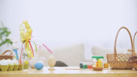 Little-boy-is-hiding-under-the-wooden-table,-full-of-Easter-decorations:-basket,-yellow-chicken,-colorful-eggs-pains-and-paint-brush.-Boy-is-playing-with-a-cute,-soft-white-bunny-with-pink-ears,-on-the-table.