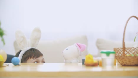 Young-adorable-boy-is-hiding-under-the-table-full-of-Easter-decorations-and-is-playing-with-Easter-bunny-in-his-hand.-Bunny-theatre.-Discussion.-Conversation:-cheerful-boy-and-a-soft-bunny.