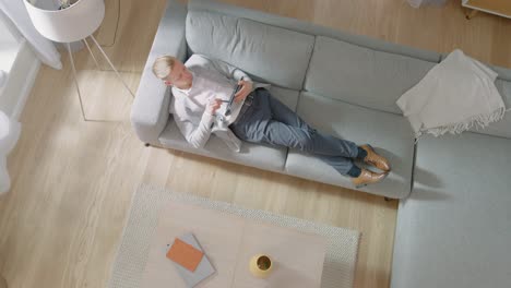 Young-Man-is-Lying-on-a-Sofa,-Working-or-Sketcing-on-a-Tablet-Computer.-Cozy-Living-Room-with-Modern-Interior,-Grey-Sofa-and-Wooden-Flooring.-Top-View-Camera-Footage.