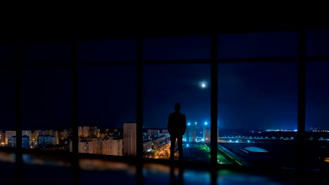 The-man-standing-near-the-window-on-the-night-city-background.-time-lapse