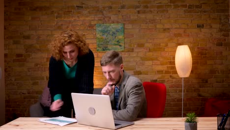 Closeup-shoot-of-young-caucasian-businessman-working-on-the-laptop-and-discussing-graphs-with-a-female-employee.-Businesswoman-using-the-tablet-sitting-on-the-couch-on-the-background